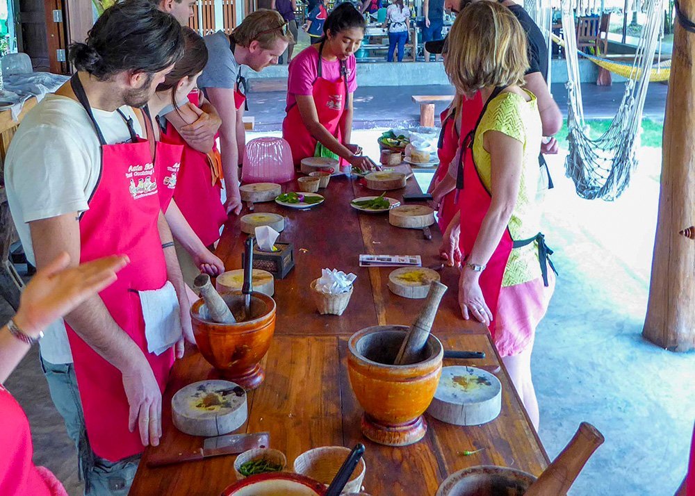 A group of people wearing red aprons participate in a cooking class, with various cooking utensils and ingredients laid out on a wooden table.