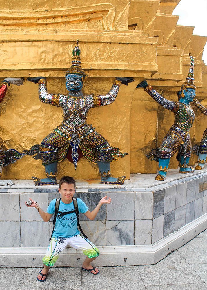 A young boy poses playfully in front of intricate, colorful guardian statues at a golden temple.