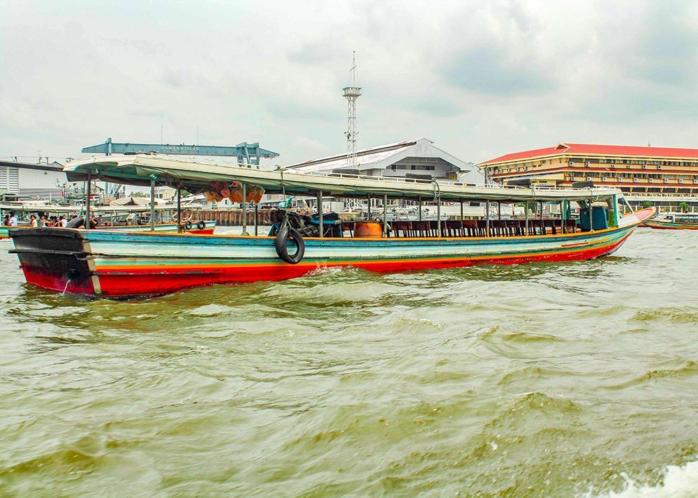 A long, colorful boat called a khlong with a green roof sails on a bustling river, with a busy dock and buildings in the background.