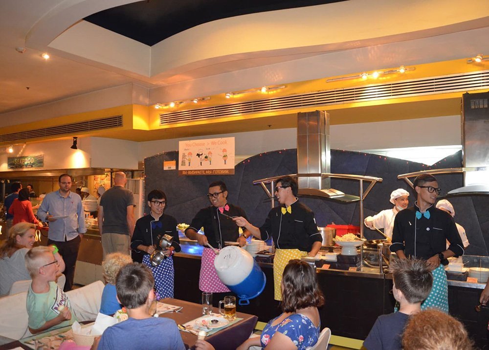 Waiters in a lively restaurant with open kitchen engage with diners, while people enjoy their meals at the tables.