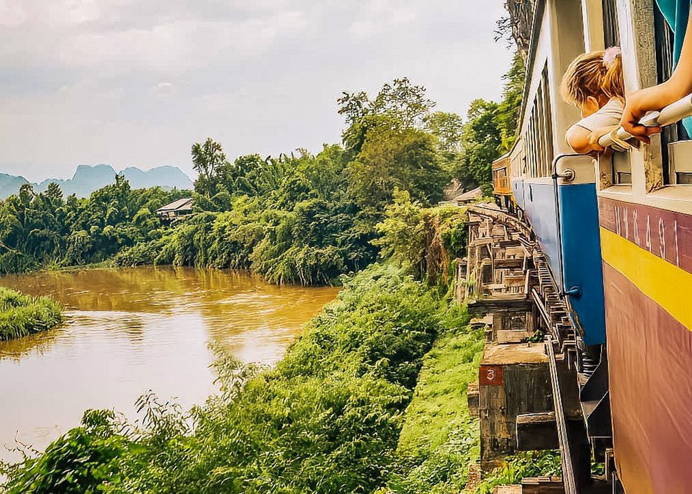 Scenic view from a train riding along the River Kwai, surrounded by lush greenery and mountains.