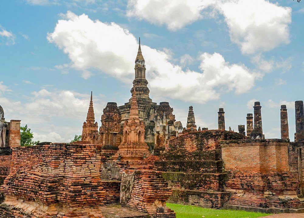 Ancient brick ruins and tall spires of a temple in Sukhothai, Thailand, set against a partly cloudy sky.