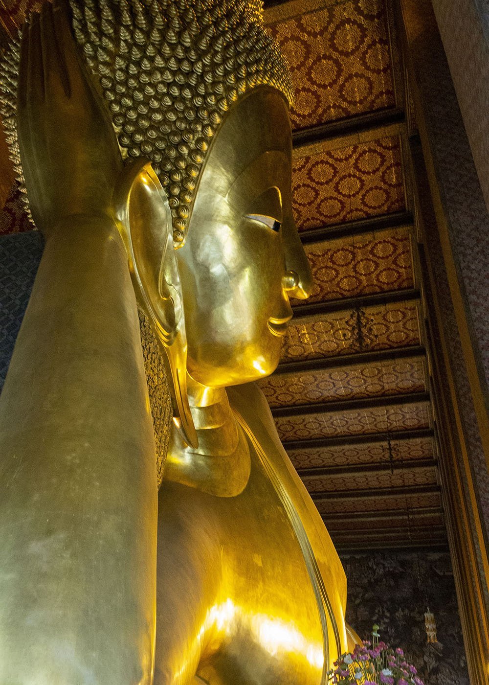 The reclining Buddha statue at Wat Pho, gleaming in gold, fills the frame. The statue is housed in an ornately decorated hall with a richly patterned ceiling.