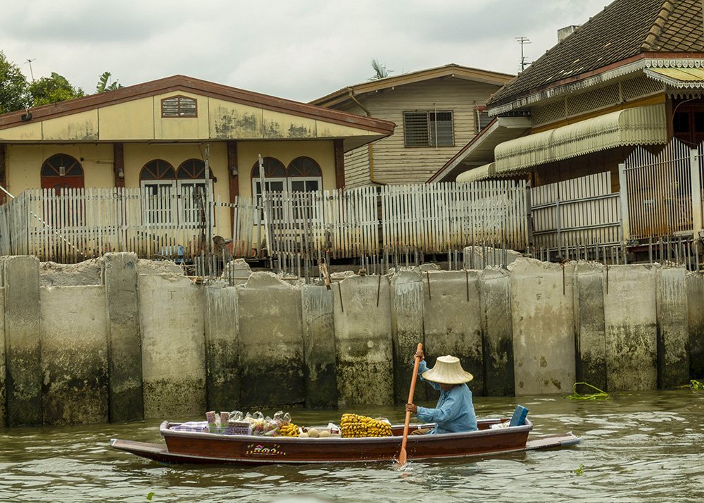 A person in a small wooden boat paddles along a canal in front of a house with a picket fence. The house has a weathered exterior, and the canal is calm.