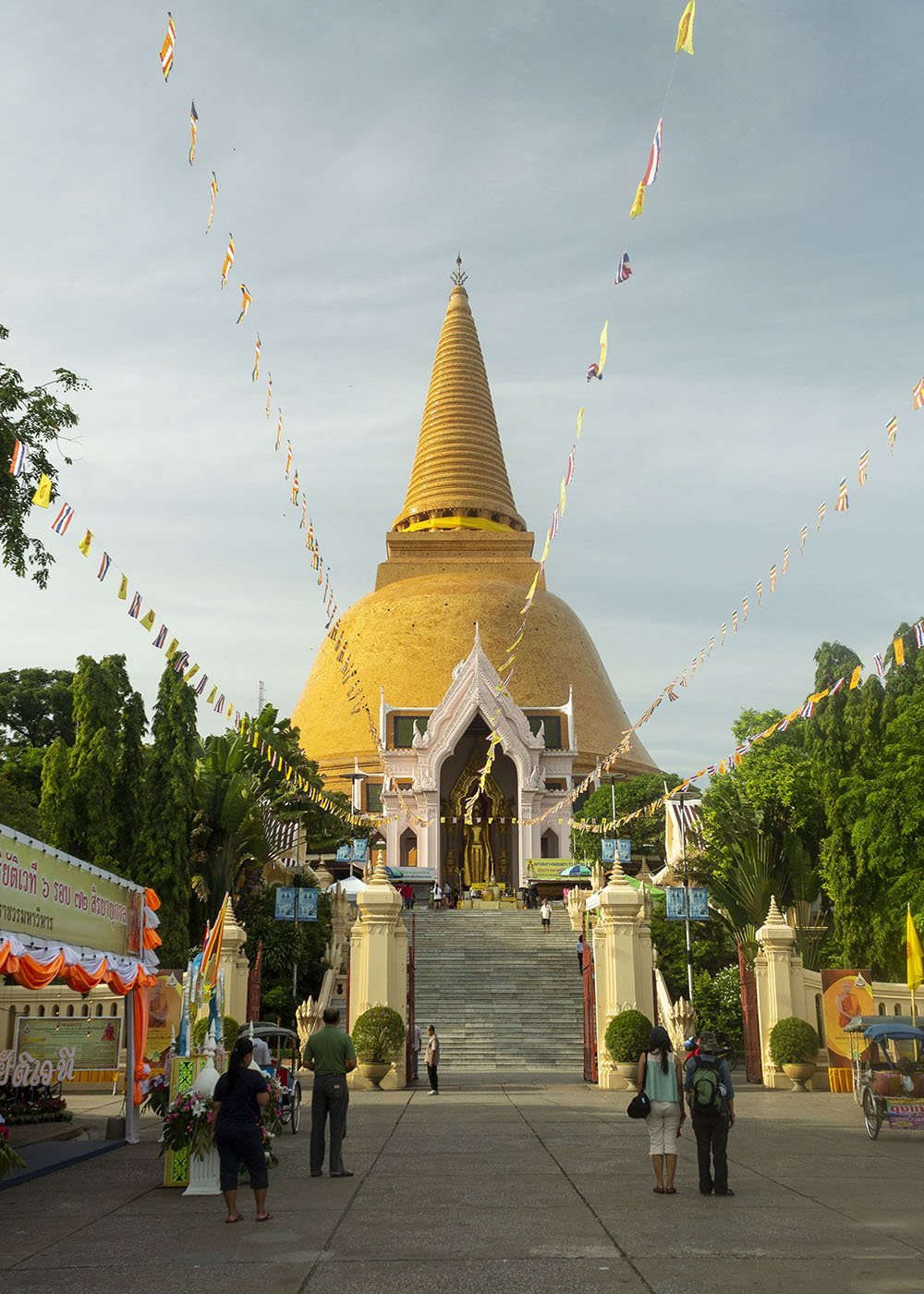 A large golden stupa with colorful flags strung around it, standing tall under a clear sky in Nakhon Pathom. People are walking up the stairs leading to the stupa, adding to the festive ambiance.