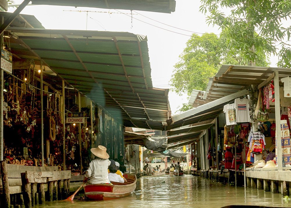 A view of a narrow canal lined with market stalls selling various goods. A person in a small boat paddles through the water, surrounded by merchandise hanging from the stalls.