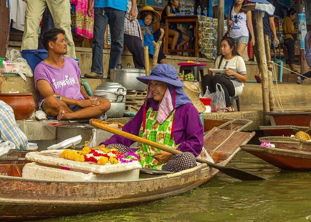 A woman wearing a hat and apron sells colorful flower garlands from her wooden boat at a floating market. Other vendors and customers are visible in the background, creating a lively atmosphere.