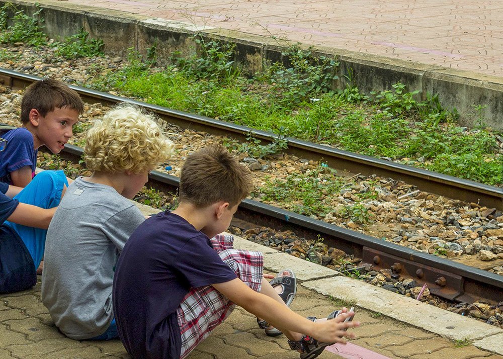 Three boys sit close to train tracks, engrossed in conversation and watching something on the ground. The scene is set at at the Kanchanaburi train station with greenery around the tracks.