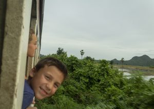 Two children lean out of a train window, smiling and looking at the lush green landscape passing by. The background features hills and trees under a cloudy sky.