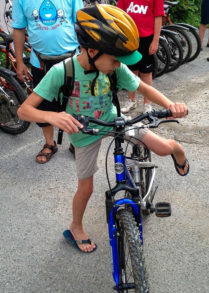 A boy wearing a yellow helmet and a green shirt mounts a bicycle, preparing to ride, with other cyclists around him.