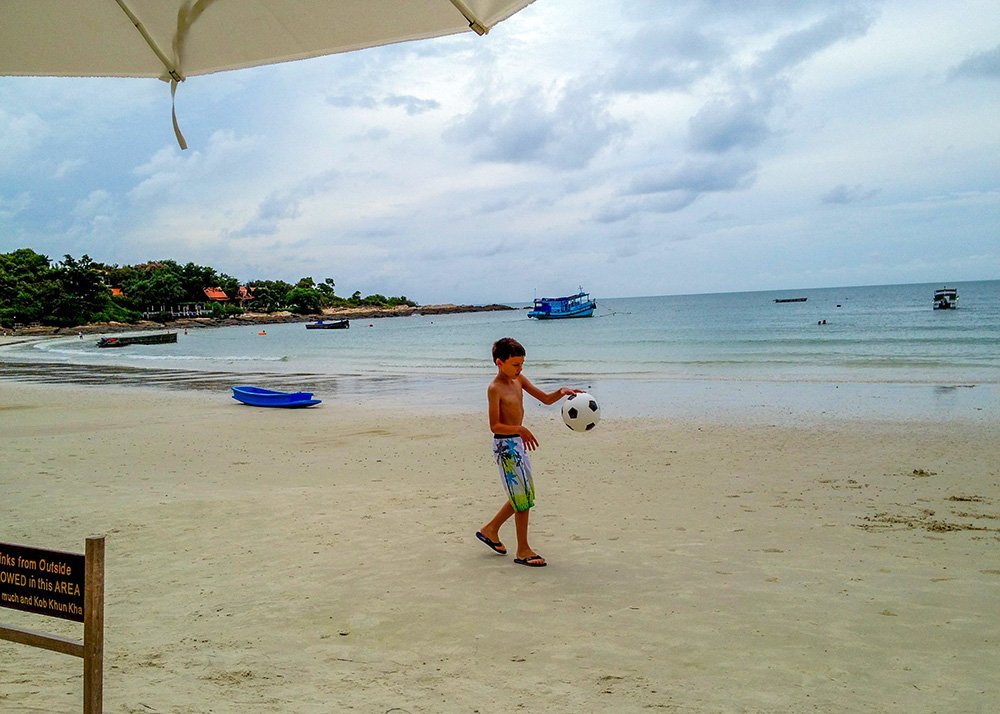 A young boy plays with a football on a sandy beach under an overcast sky. The calm sea and several small boats are in the background, with a few people swimming in the distance.