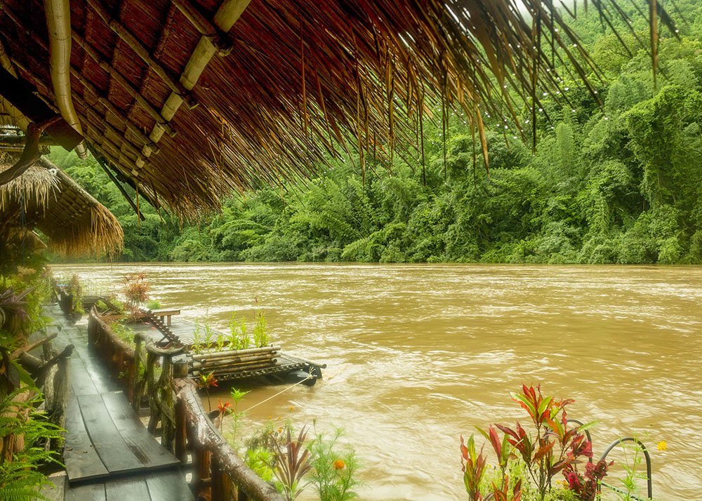 A wooden walkway with a thatched roof overlooks a muddy river surrounded by lush, dense jungle. Several small platforms with plants are tied to the side, adding a touch of greenery to the rustic setting.