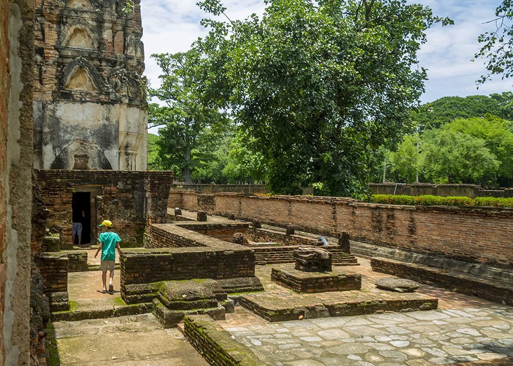 A boy walks through the ancient ruins of a temple complex in Sukhothai, with old brick structures and large trees providing a sense of history and tranquility.