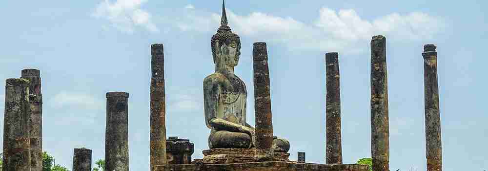 A large, ancient Buddha statue sits among tall, weathered columns at Sukhothai Historical Park. The sky is partly cloudy, adding to the majestic atmosphere of the ruins.
