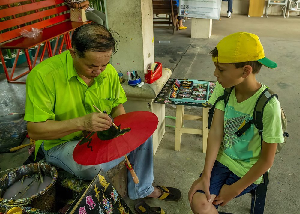 A local artisan paints a design on a red umbrella while a young boy sitting next to him watches attentively. The artisan's workspace is filled with paints and tools.