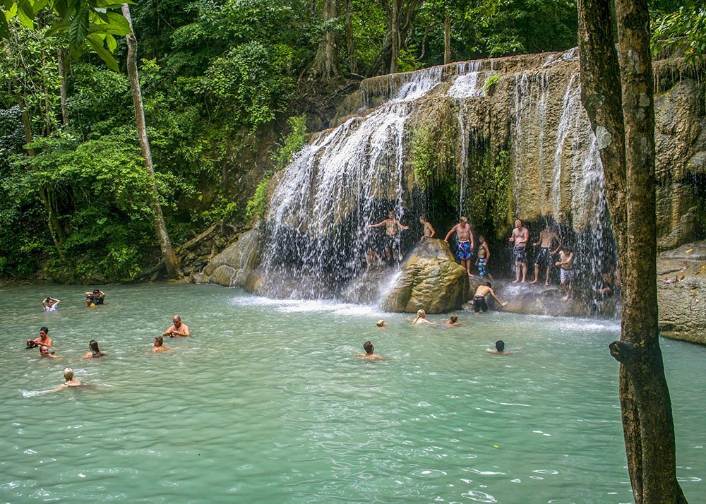 People swim and relax in a natural pool at the base of a cascading waterfall in a forested area. The waterfall is surrounded by greenery, creating a serene and refreshing atmosphere. This is one of the Erawan waterfalls in Thailand.