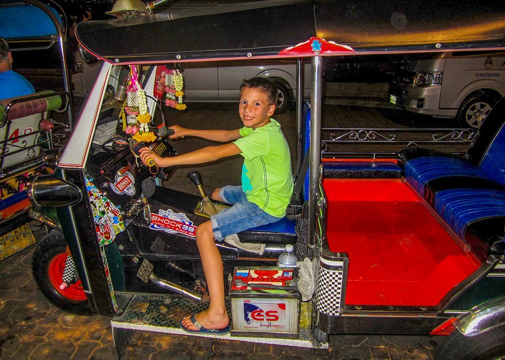 A young boy sits smiling in the driver's seat of a brightly decorated tuk-tuk at night. The tuk-tuk has colorful garlands hanging from the roof and vibrant seats.