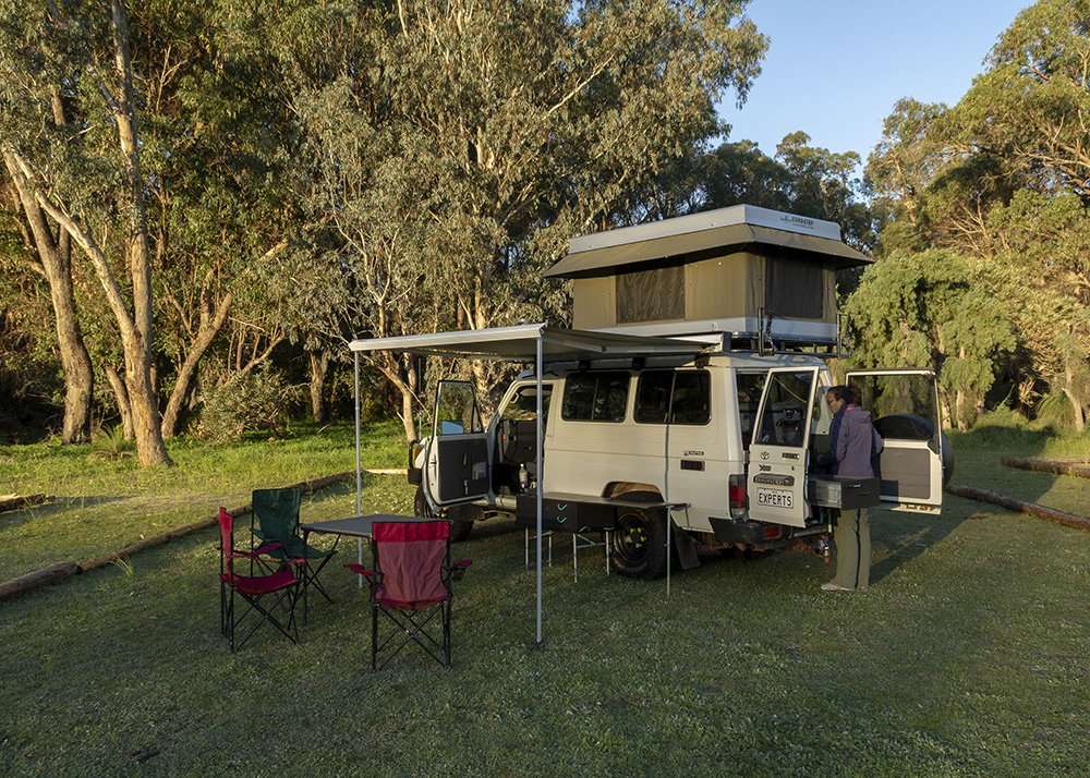 A camper van with a rooftop tent set up in a grassy campsite, surrounded by tall trees and camping chairs.
