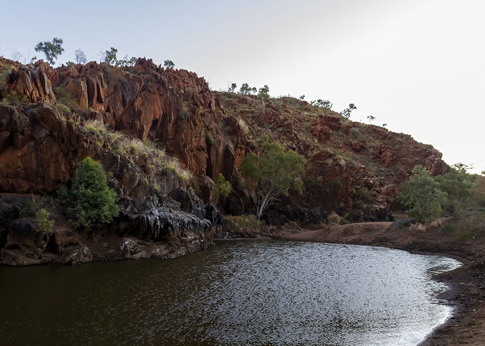 A tranquil natural pool surrounded by rocky red cliffs, with green vegetation along the banks under an overcast sky.