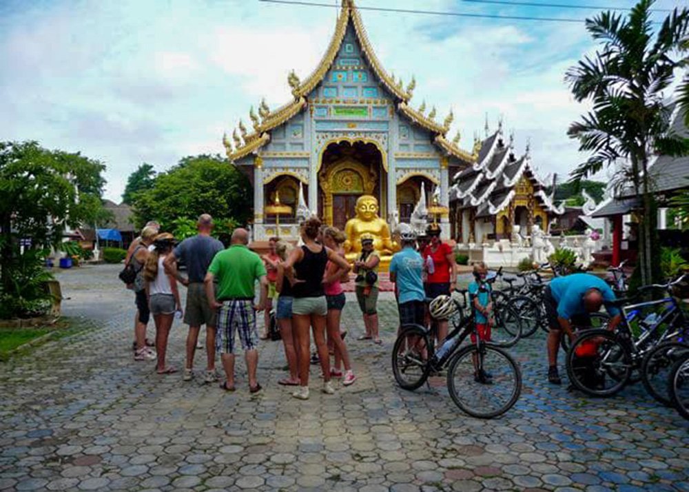 A group of tourists stands in front of an ornate temple with a golden Buddha statue and intricate architectural details. Bicycles are parked nearby, suggesting they arrived on a bike tour.