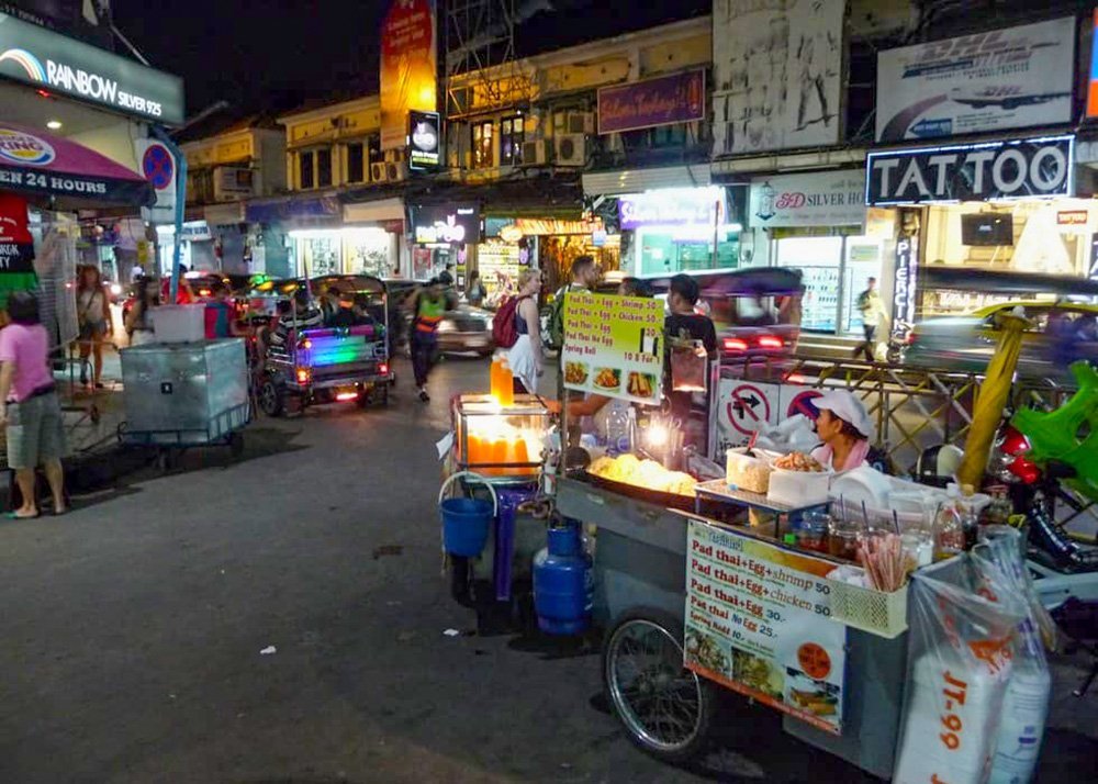 A busy night market street with food vendors, tuk tuks, and a variety of illuminated signs and storefronts.