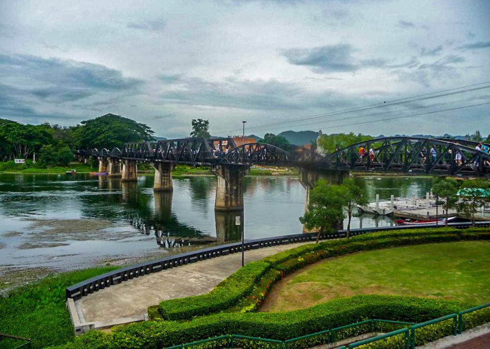 The famous Bridge on the River Kwai spans across a calm river, with lush greenery and hills in the background. The bridge is a historic structure, with a path leading up to it.
