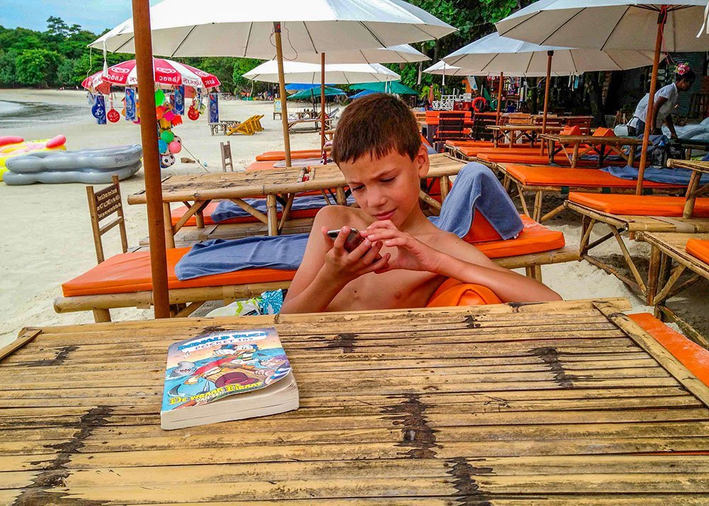 Boy sitting at a bamboo table on a beach, looking at a phone, with a book and beach umbrellas in the background.