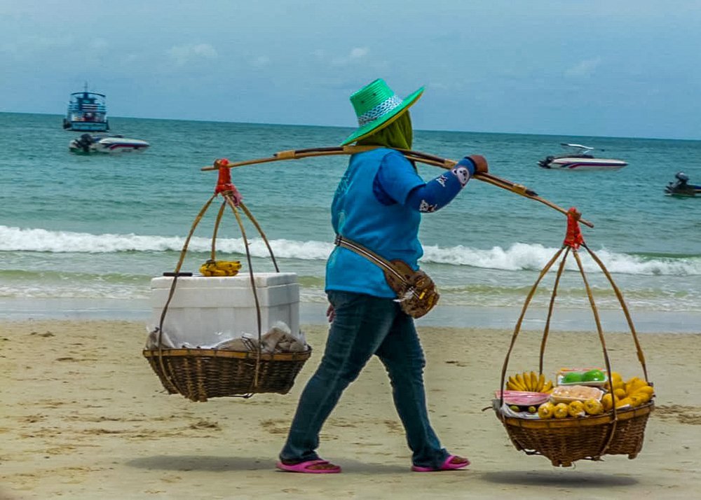 Vendor carrying goods in baskets on a yoke, walking along the beach in Koh Samet, Thailand.