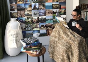 a woman sits in a chair with a cup of coffee in hand and a brown blanket on her lap. She's reading The Ultimate Travelist from Lonely Planet. Next to here is an small table with a few travel related coffee table books.