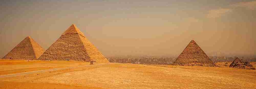 Panoramic view of three pyramids at Giza, including the Great Pyramid, with a vast desert landscape in the foreground.