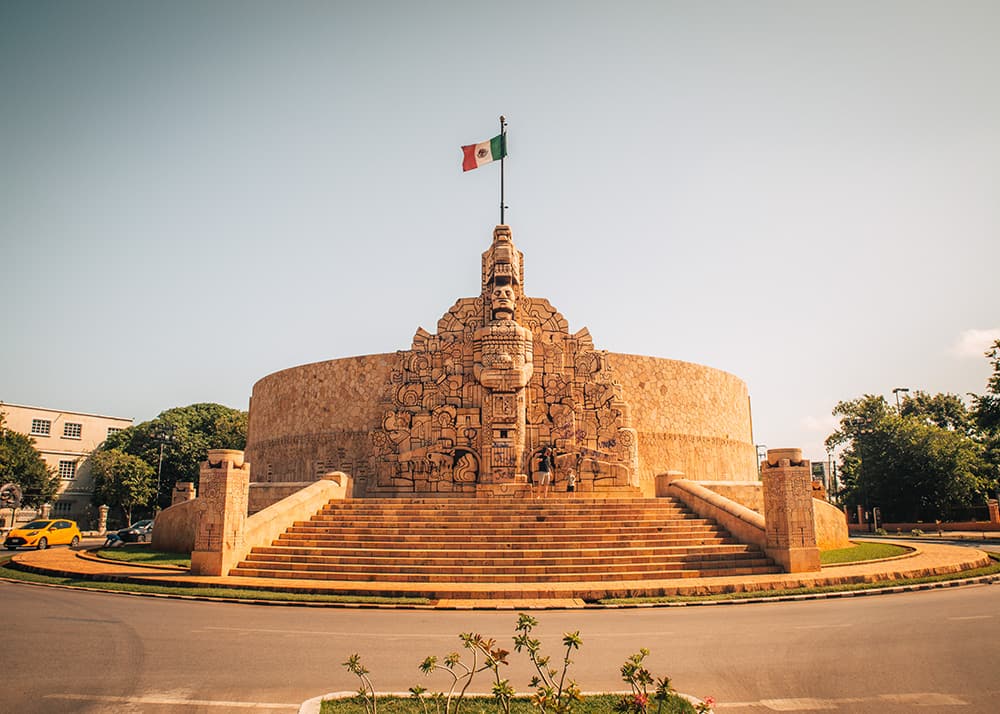 The Monumento a la Patria in Mérida, Mexico, featuring detailed carvings and a Mexican flag on top. The weather is clear and sunny, highlighting the monument's intricate design and historical significance.