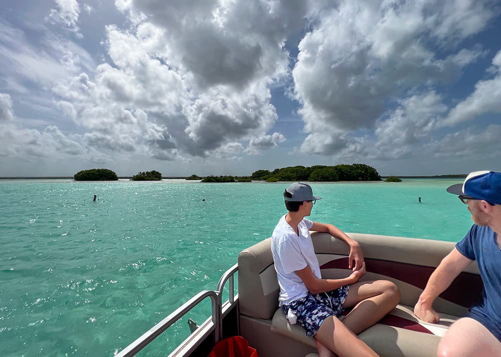 Two people sit on a boat in Bacalar Lagoon, Mexico, enjoying the view of the turquoise water and distant green islands under a partly cloudy sky. The relaxed atmosphere highlights the natural beauty of Bacalar.