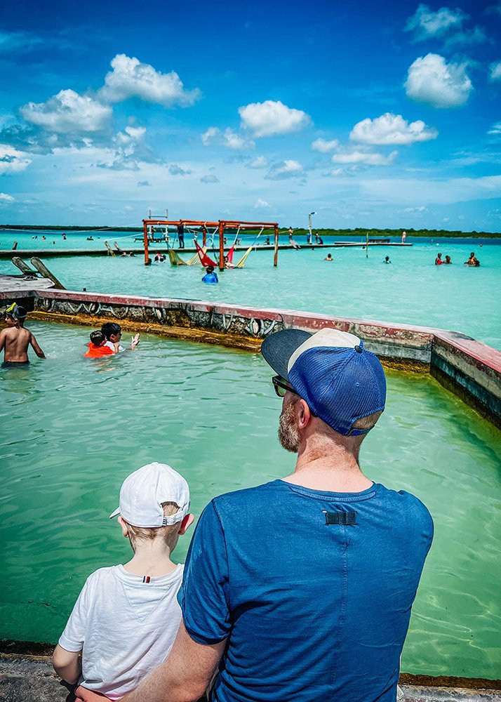 A group of people, including children and adults, are seated and standing by the clear, turquoise waters of Lake Bacalar, Mexico, enjoying a sunny day. In the background, lush greenery and a calm lake enhance the serene setting.