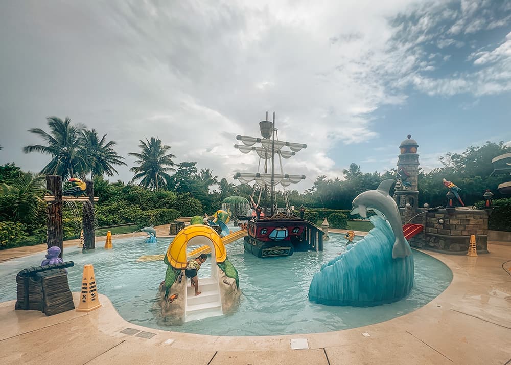 A children's pool at a beach park in Cozumel, Mexico, with kids splashing and playing in the water. The tropical weather is perfect, and the pool area is surrounded by palm trees and loungers.