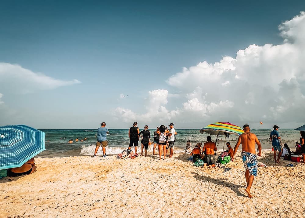 A family relaxes on the sandy beach in Playa Del Carmen, Mexico, with children playing in the sand and adults lounging under umbrellas. The crystal-clear water and sunny weather make for a perfect beach day.