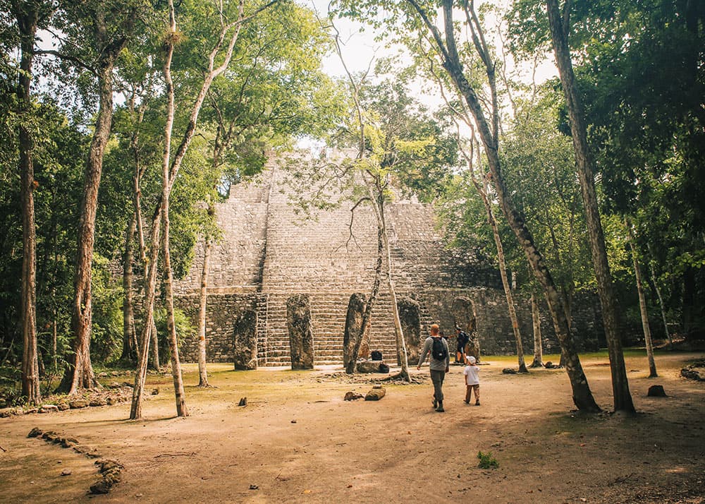 A family walks toward a large stone pyramid at the Calakmul ruins, Mexico, surrounded by dense jungle. The scene is shaded by tall trees, with dappled sunlight creating a tranquil atmosphere.