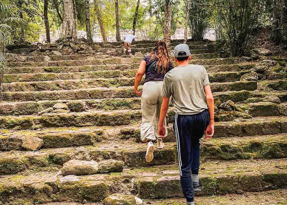 Visitors climb the moss-covered stone steps at the Calakmul ruins in Mexico, surrounded by dense forest. The overcast sky adds to the mystical and ancient feel of the location.