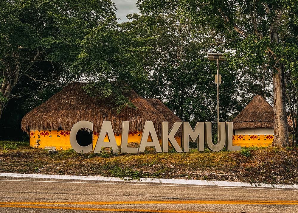 A sign reading "CALAKMUL" stands prominently in front of traditional thatched-roof buildings at the entrance of Calakmul, Mexico. The scene is lush with greenery, creating a welcoming and historical ambiance.