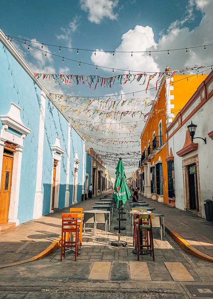 A vibrant street in Campeche, Mexico, lined with brightly colored buildings and decorated with hanging ribbons. The sunny weather and clear sky make the scene lively and festive.