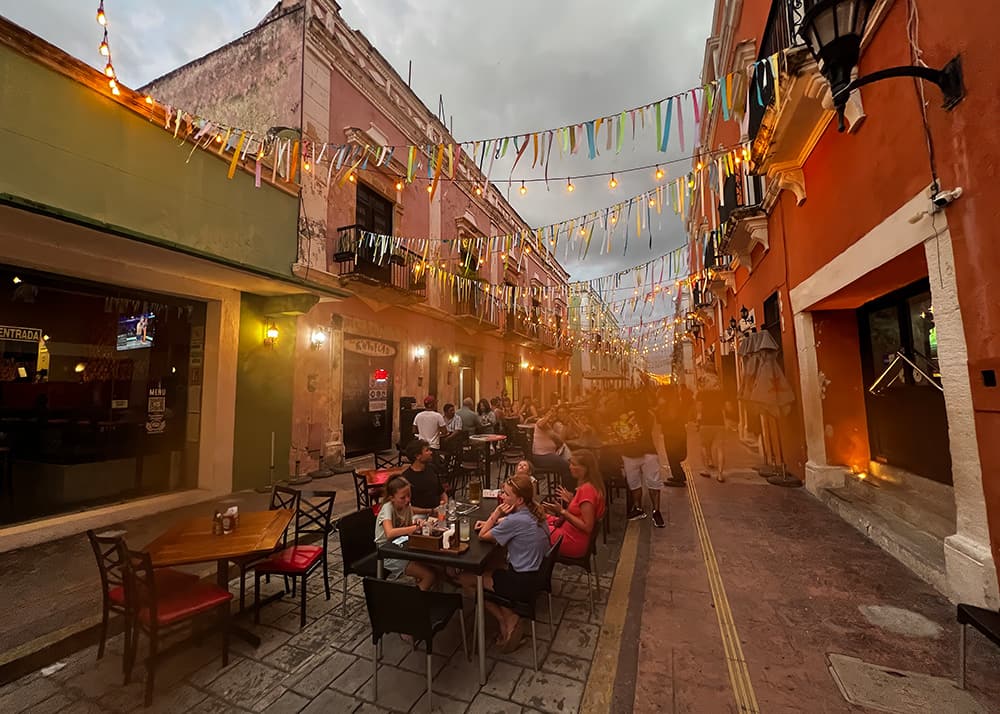People enjoy an outdoor dinner on a narrow street in Campeche, Mexico, under strings of lights as dusk falls. The warm lighting and colorful buildings create a cozy and inviting atmosphere.