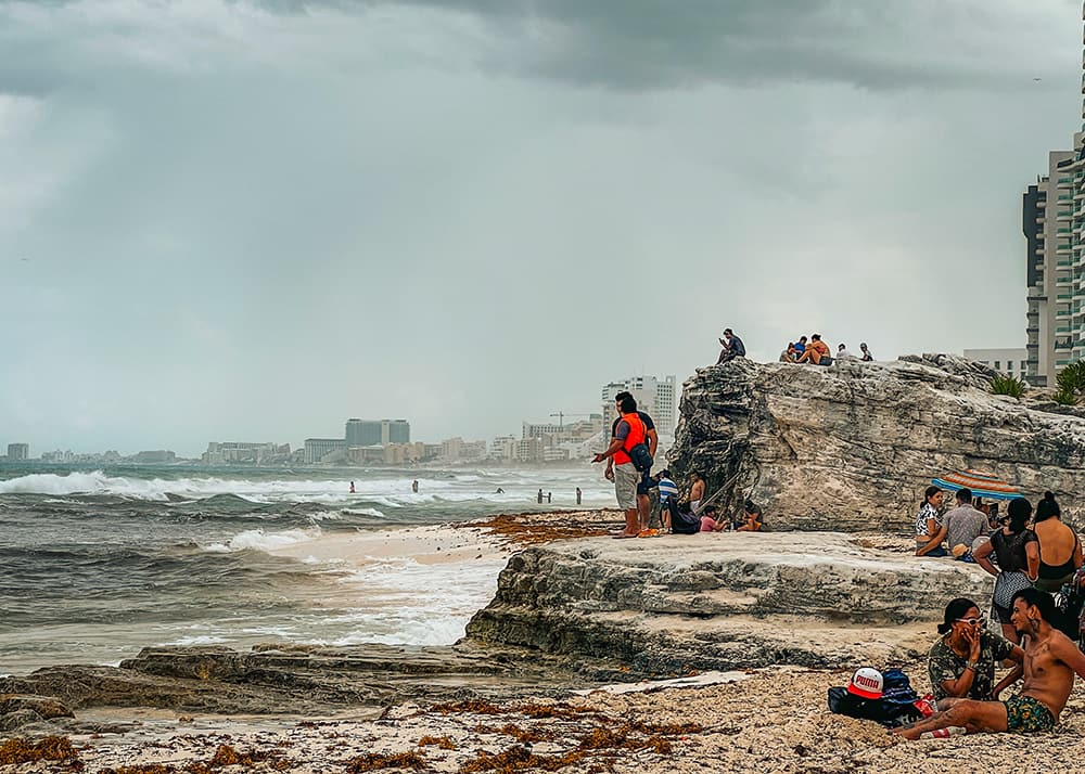 A panoramic view of the beach strip in Cancun's hotel zone, Mexico, showcasing a cliff with people standing on top of it and chilling on the white sandy beaches next to the turquoise Caribbean Sea.