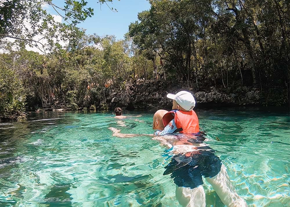 A boy with an orange life and a white cap on the back on his father in the water of a cenote. His teen son is swimming in front of him. Jungle surrounds the cenote.