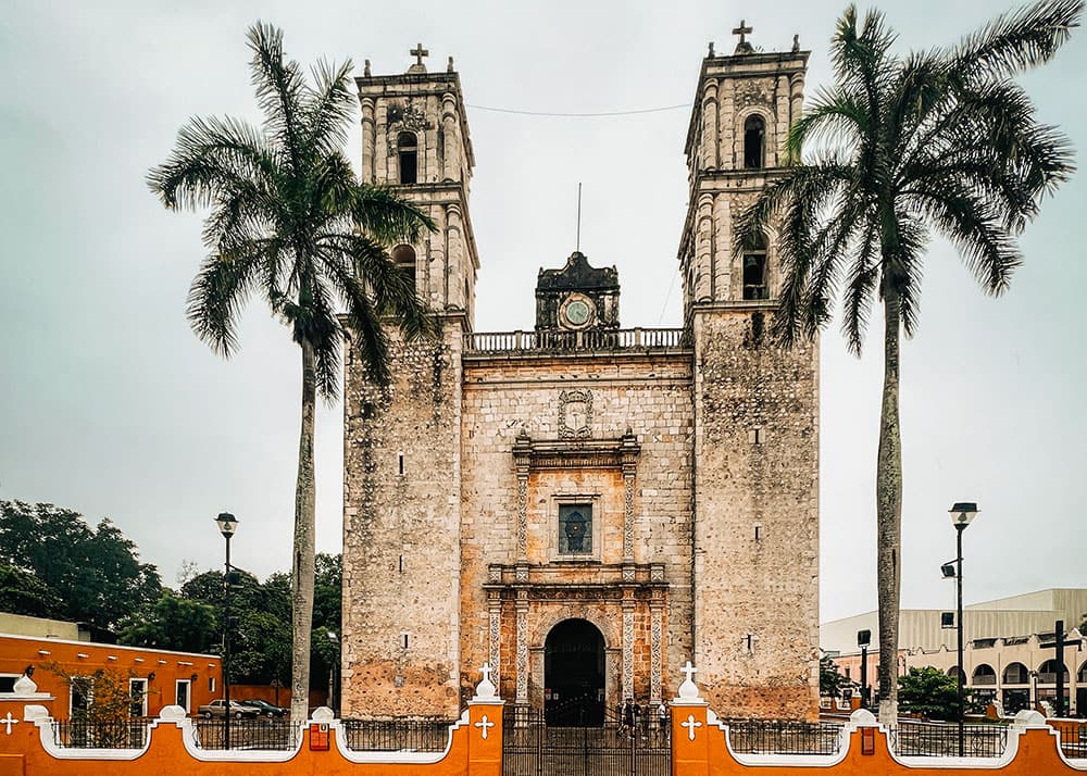 The Church of San Servacio in Valladolid, Mexico, framed by tall palm trees and featuring twin bell towers. The weather is overcast, giving the scene a calm and serene atmosphere.