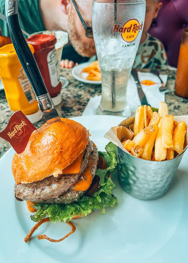 A table in Cancun's Hard Rock Cafe is filled with an empty tall glass and with a plate filled with a huge hamburger with cheese and a bucket of fries.