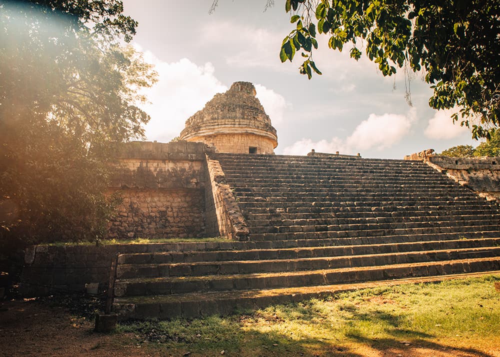 The El Caracol observatory at Chichen Itza, Mexico, with its circular structure and stepped platform. The sun filters through the trees, casting a warm glow on the ancient ruins.