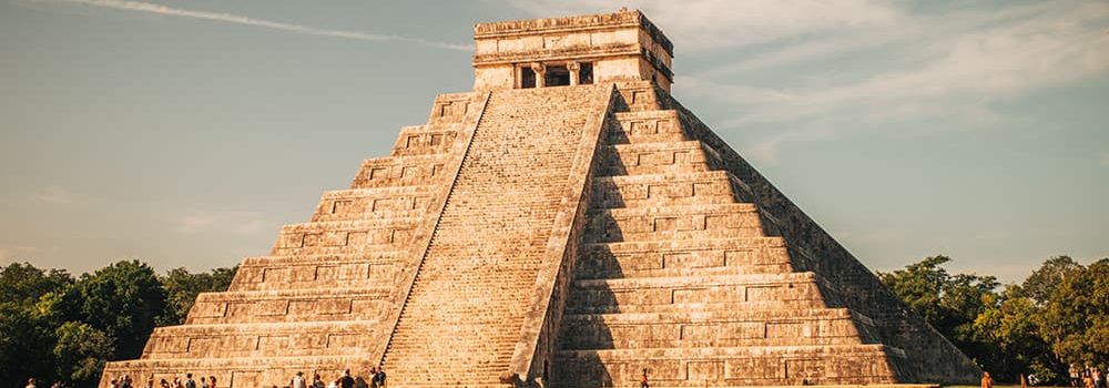 The iconic El Castillo pyramid at Chichen Itza, Mexico, stands tall under a blue sky with light clouds. Tourists gather around the base, appreciating the grandeur and historical importance of the ancient structure.