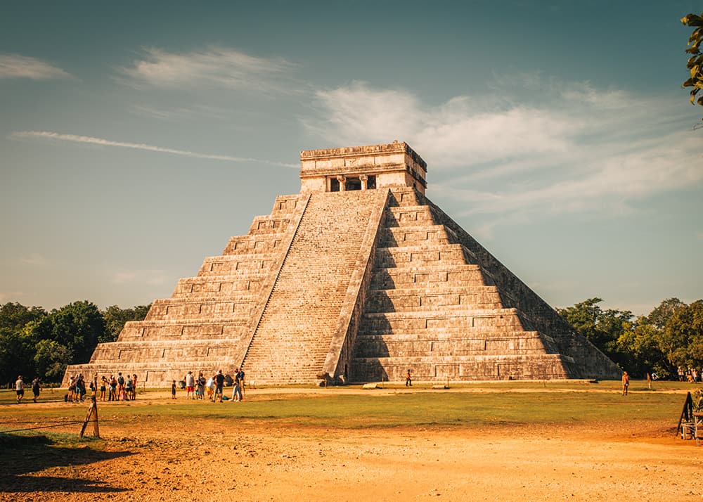 The iconic El Castillo pyramid at Chichen Itza, Mexico, stands tall under a blue sky with light clouds. Tourists gather around the base, appreciating the grandeur and historical importance of the ancient structure.
