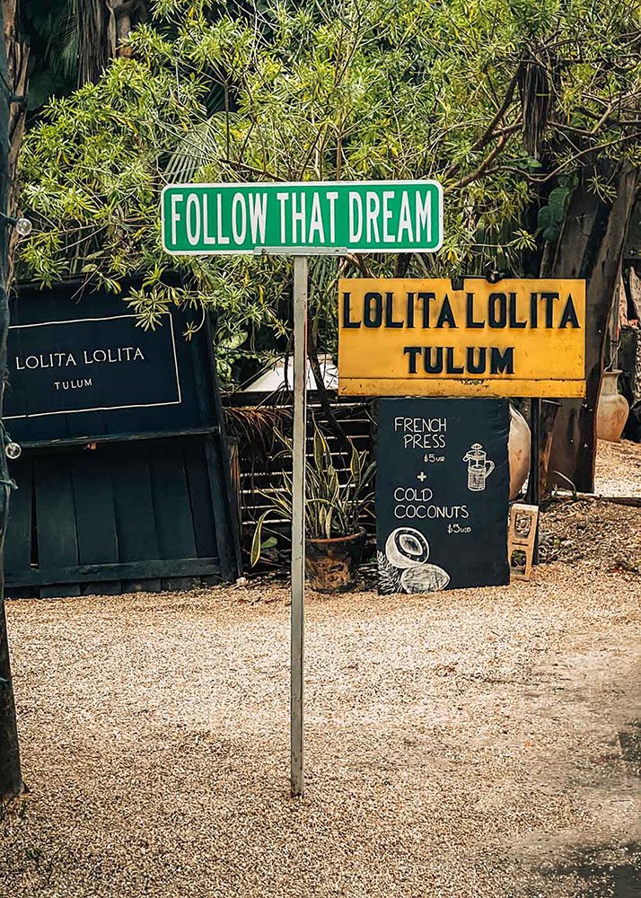 A whimsical sign reading "Follow That Dream" stands by a roadside in Tulum, Mexico. The weather is bright and sunny, with palm trees and a clear blue sky creating a tropical, motivational scene.