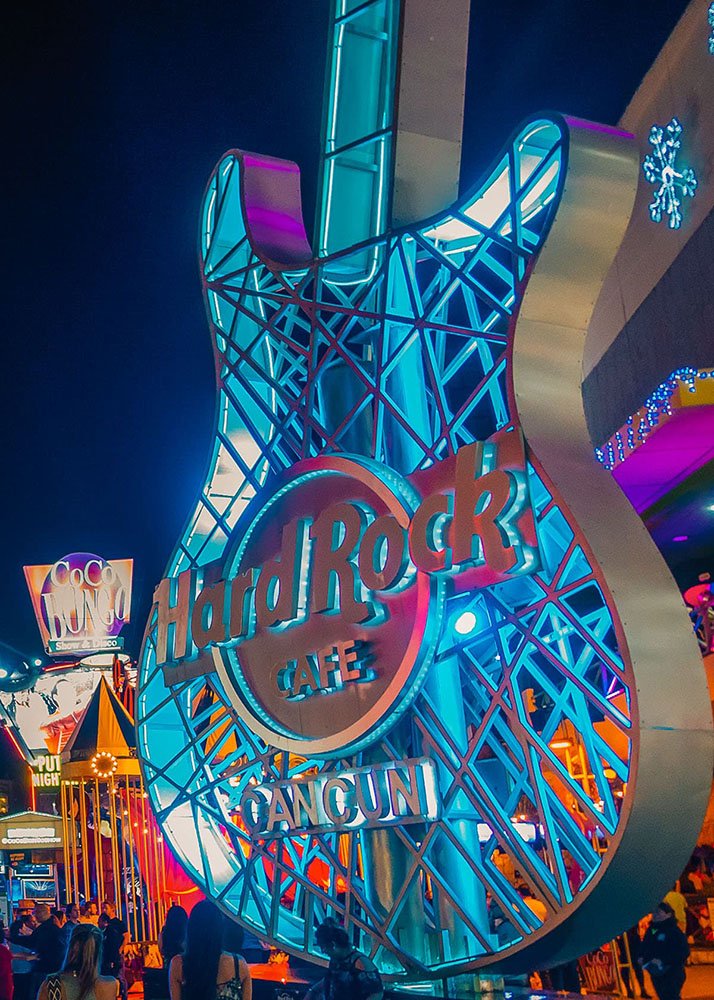 The huge Hard Rock Cafe billboard in the shape of a guitar and lit up in different hues of blue and purple stands tall next to the entrance of the restaurant.