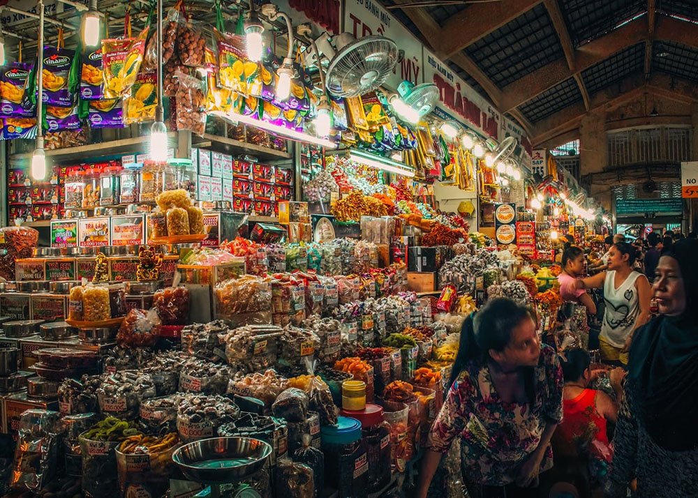 A stall in the HCMC night market is filled with all sorts of herbs and medicine. A woman is selling something to another woman, while tourists walk by and enjoy the market.